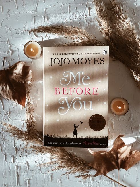 A flat-lay photograph features a paperback copy of the novel 'Me Before You' by Jojo Moyes placed on a textured white brick surface. The book’s cover prominently displays the title in light blue and pink text, with the author's name above in black. Surrounding the book are decorative elements including two small, round, beige candles in shallow holders, dry pampas grass arranged in an arc around the book, a dried brown leaf, and a dried flower with brown petals. The scene is well-lit with soft, natural light creating gentle shadows, emphasizing the arrangement's detail and texture. This setting highlights elements of home decor, reading, and peaceful moments that could occur during or after a house move, aligning with themes of home relocation and packing preparation, as seen in removals services like those by Wimbledon Man and Van.