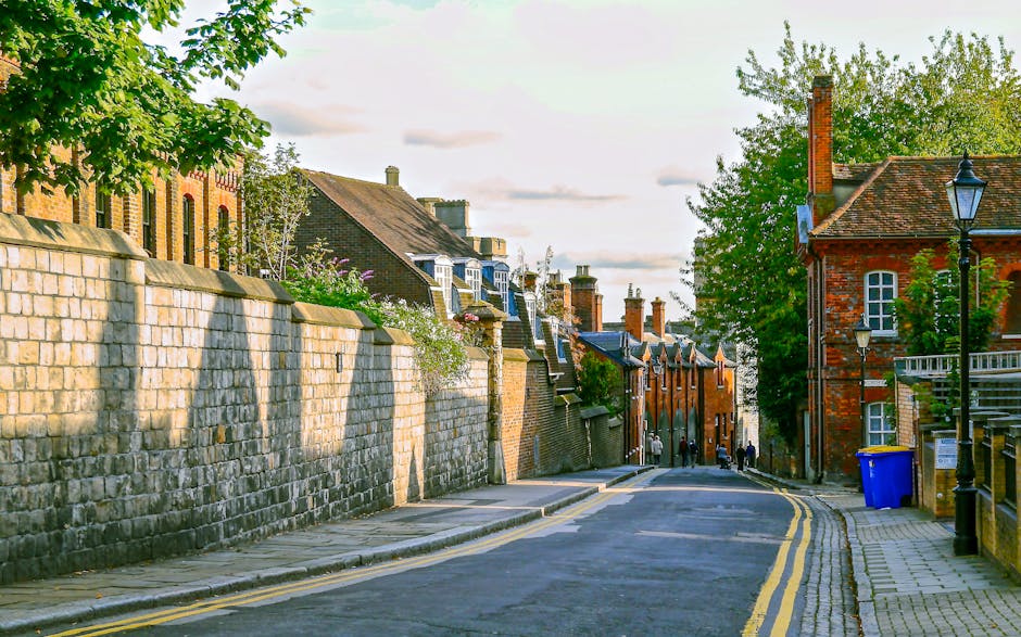 A quiet residential street in Wimbledon featuring a paved tarmac road with two yellow lines along the curb, flanked by a high stone wall on the left side and a row of brick houses with chimneys and slate roofs on the right. The sidewalk consists of grey paving slabs, with a black lamppost and a blue waste bin visible on the right. Several people are seen walking in the distance, and lush green trees along with a partly cloudy sky provide natural lighting, indicating daylight. This scene captures a typical area where house removals and furniture transport are undertaken, with the environment conducive to loading and moving activities, reflecting the context of home relocation services like those offered by Wimbledon Man and Van.