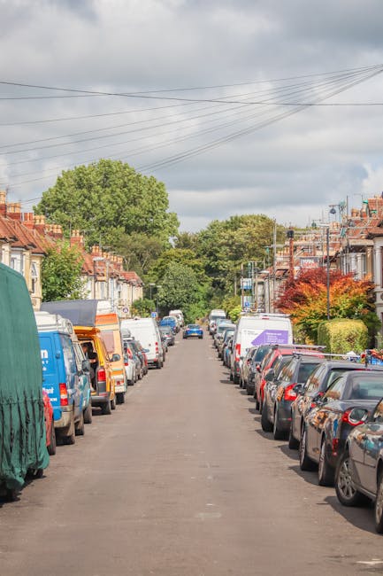 A narrow cobbled street in Wimbledon Village with tall brick terraced houses on the right side, featuring large windows and white window frames. Potted plants and small shrubs line the front of the houses, some placed on the pavement, while a large leafy tree extends its branches over the street from the left. The sky overhead is partly cloudy with patches of blue visible. In this image, there are no visible objects or furniture related to house removals or moving processes, but it depicts the residential environment where home relocation services by Wimbledon Man and Van could take place, emphasizing the tight streets often encountered during furniture transport and packing and moving activities.